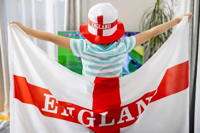 A boy holding England flag and watching soccer game on TV at home