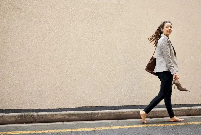 Woman in street, walking at wall with mockup and barefoot, relax and travel for work holiday in city. Space, road and freedom, person on walk with shoes in hands, morning commute and business trip.