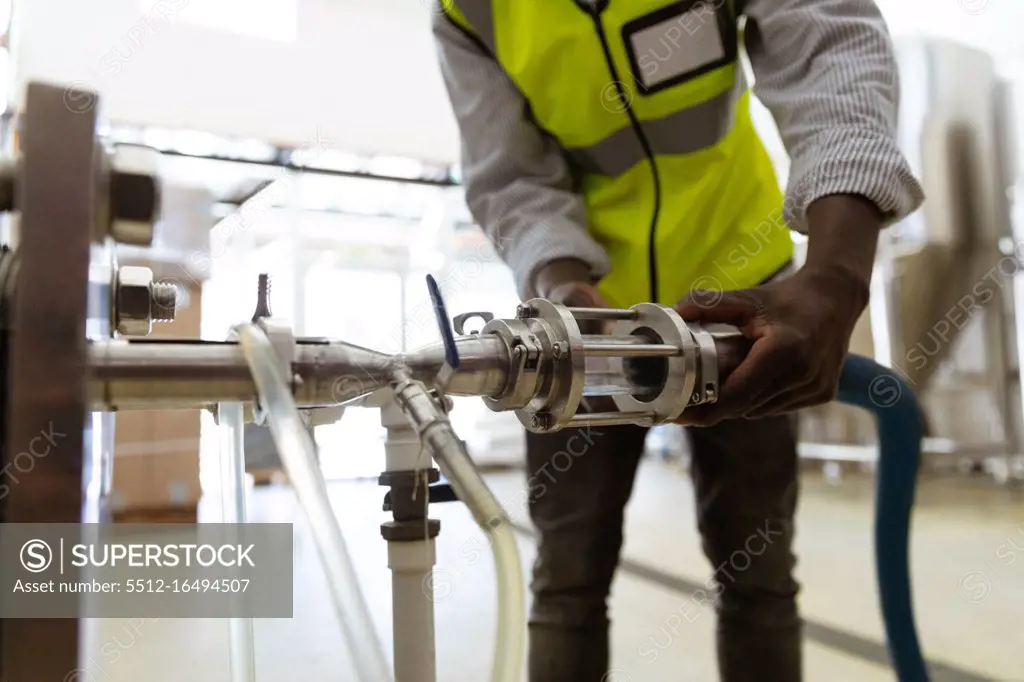 Mid section view of an African American man working at a microbrewery, wearing a high visibility vest, inspecting a tank pipe.