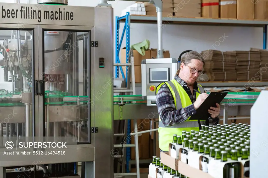 Caucasian man wearing high visibility vest, working in a microbrewery, holding a file and writing data while checking bottles of beer prepared for delivery..