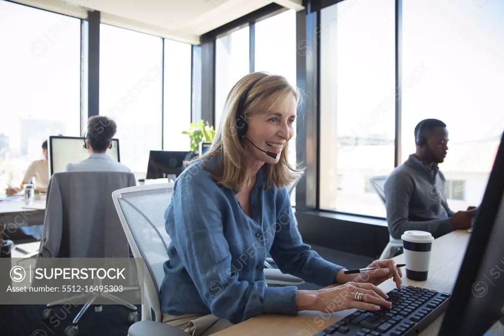 A Caucasian businesswoman working in a modern office, sitting at a desk, using a laptop computer, wearing headset and talking, with her colleagues working in the background