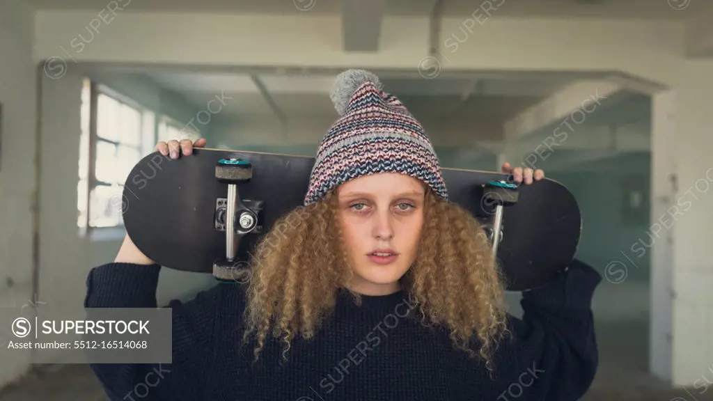 Front view of a young Caucasian woman with curly hair wearing a black long sleeves and beanie while holding a skateboard overshoulder and looking intently at the camera inside an empty warehouse