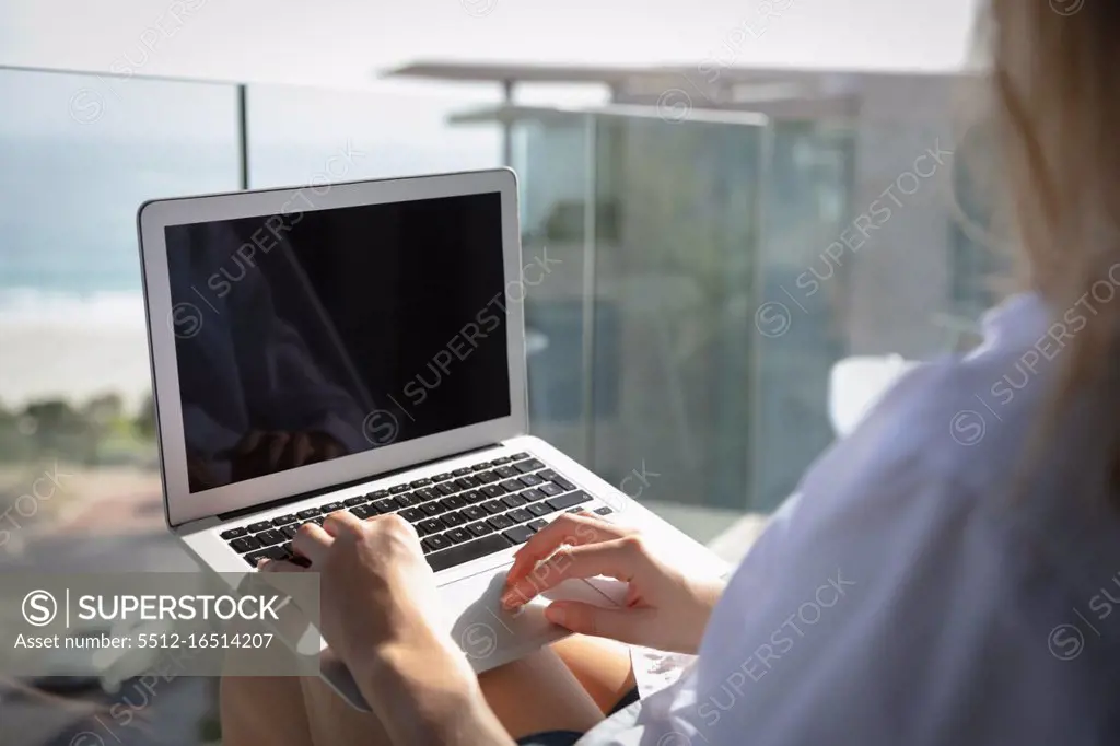 Over the shoulder view of a young Caucasian woman wearing a white shirt, sitting on a balcony using a laptop, seafront and buildings in the background. She is relaxing and practicing selfcare.