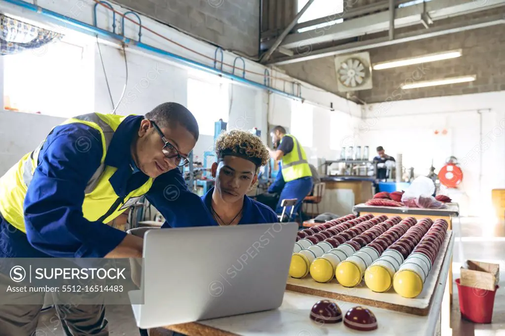 Front view close up of a young mixed race male manager standing and looking at a laptop with a young mixed race male worker sitting at a workbench with finished cricket balls at a cricket ball factory. They are working in a clothing factory.