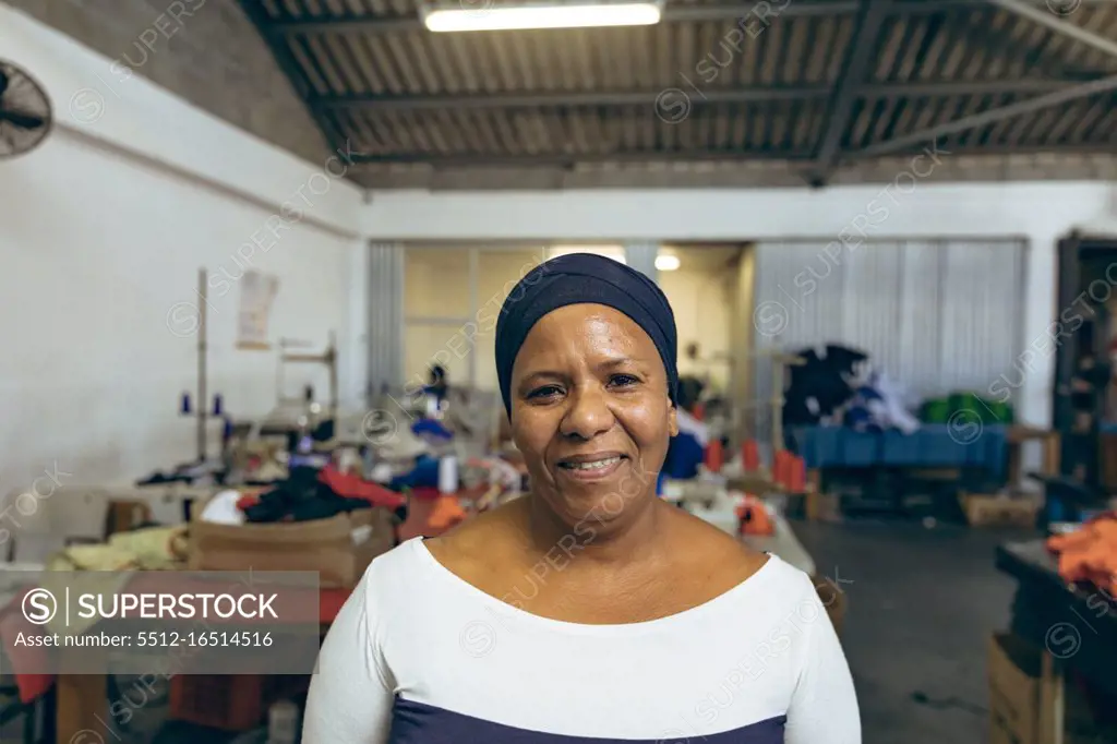 Portrait close up of a middle aged mixed race woman in a brightly lit sports clothing factory, looking to camera and smiling. They are working in a clothing factory.