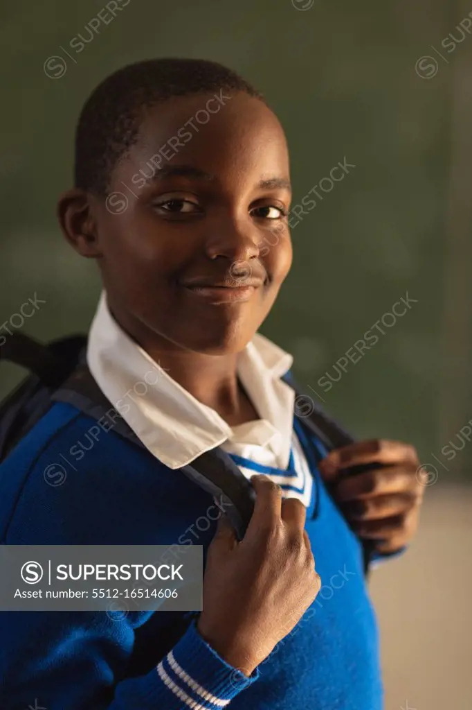 Portrait close up of a young African schoolboy wearing his school uniform and schoolbag, looking straight to camera smiling, at a township elementary school