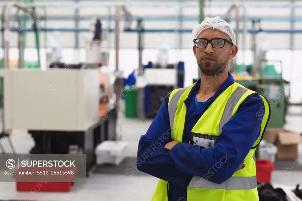 Portrait close up of a middle aged mixed race male factory worker wearing glasses and workwear looking to camera with arms crossed in a warehouse at a processing plant