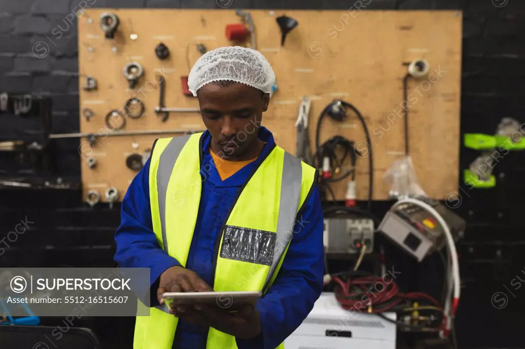 Front view close up of a young African American male factory worker using a tablet computer in the machine shop at a processing plant, with equipment and tools in the background