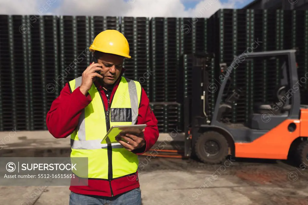 Front view close up of a middle aged Caucasian male factory worker on the phone using a tablet computer beside a forklift truck and stacks of pallets outside a warehouse at a factory 