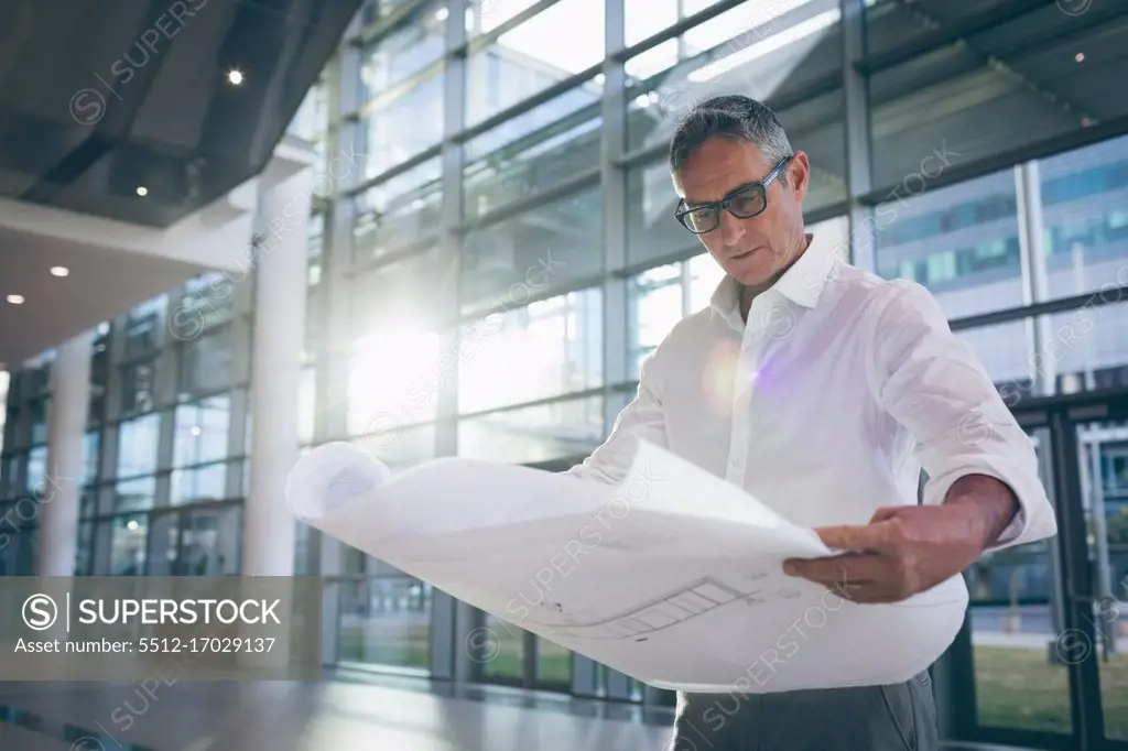 Front-view of a concentrated businessman reading a blueprint plan in office next to big windows showing the city in the background