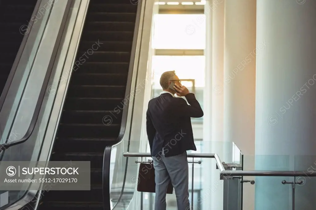 Rear view of businessman with briefcase talking on the phone near the escalator in office