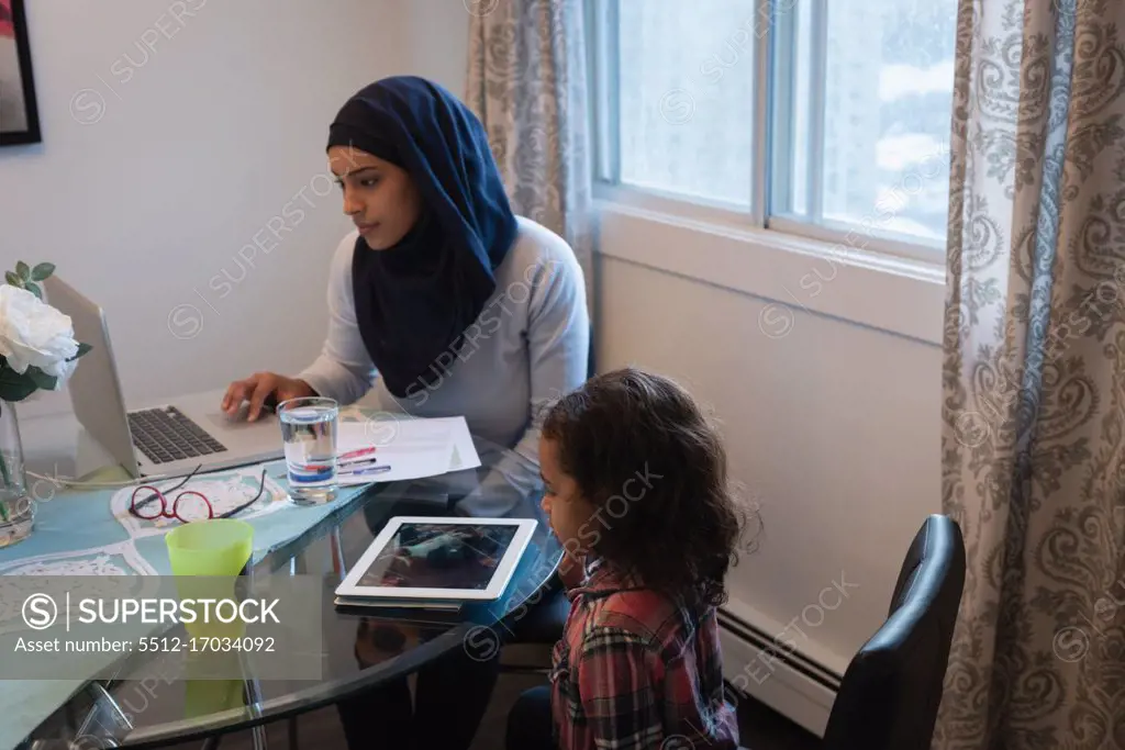 Side view of mixed race mother wearing hijab using laptop while daughter looking at digital tablet at home. They are sitting around a table in living room