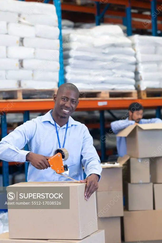 Portrait close-up of happy mature African-american male staff packing cardboard box with tape gun dispenser in warehouse. Asian male worker is unpacking cardboard box in the background. This is a freight transportation and distribution warehouse. Industrial and industrial workers concept