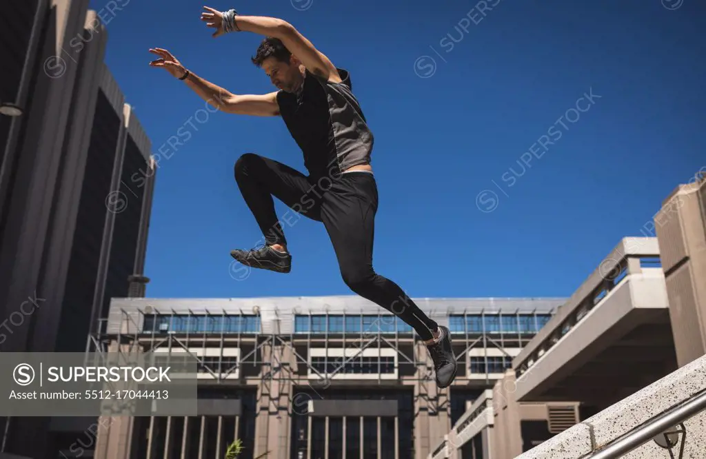 Side view of a Caucasian man practicing parkour by the building in a city on a sunny day, jumping on concrete handrail.