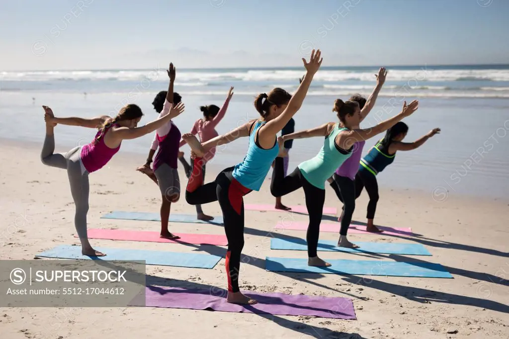 Side view of a multi-ethnic group of female friends enjoying exercising on a beach on a sunny day, practicing yoga, standing in yoga position, stretching..