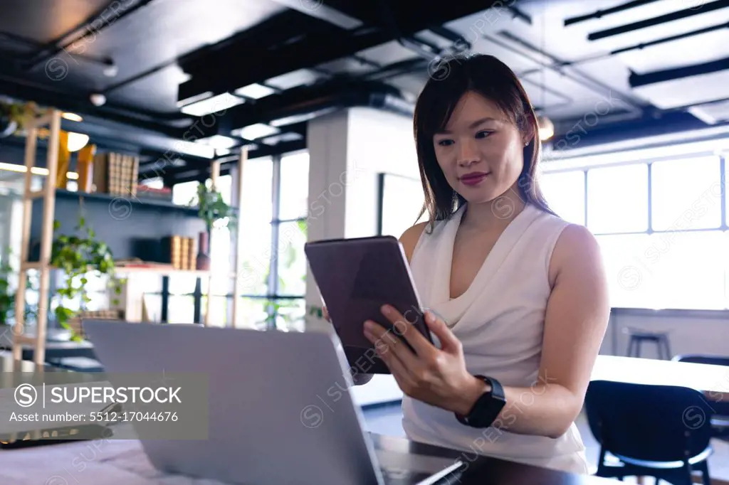 Front view of an Asian businesswoman wearing smart clothes, working in the modern office, standing by a desk and using her laptop computer and tablet. 