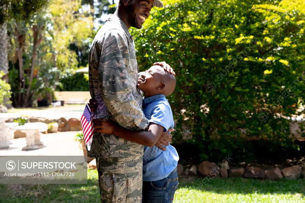 African American male solider wearing uniform embracing his son holding a USA flag, standing in their garden on a sunny day, smiling and interacting.