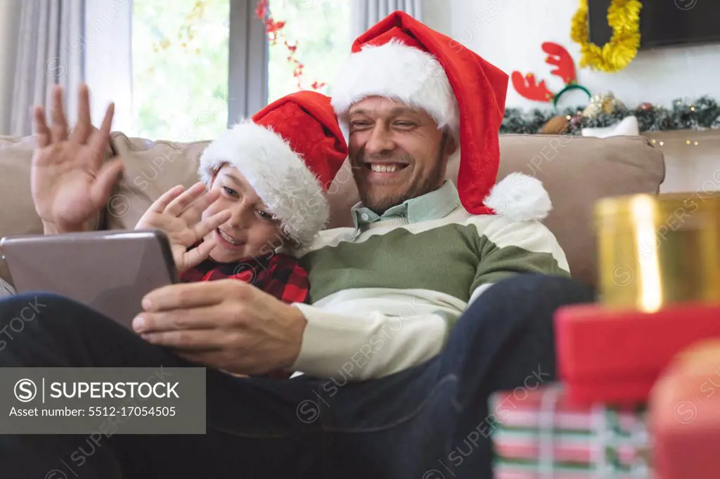 Caucasian man at home with his son at Christmas, wearing Santa hats sitting on sofa in living room, using digital tablet, waving. Social distancing during Covid 19 Coronavirus quarantine lockdown.