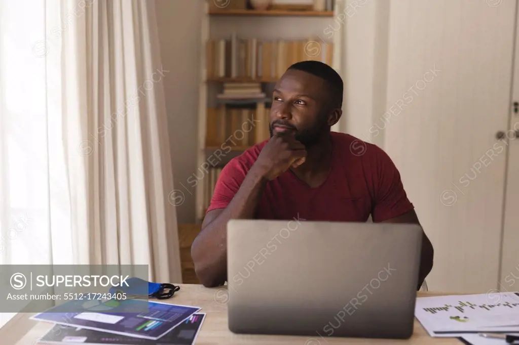 Thoughtful  african american man looking out of window with laptop, face mask and documents on table at home. social distancing during covid 19 coronavirus quarantine lockdown.