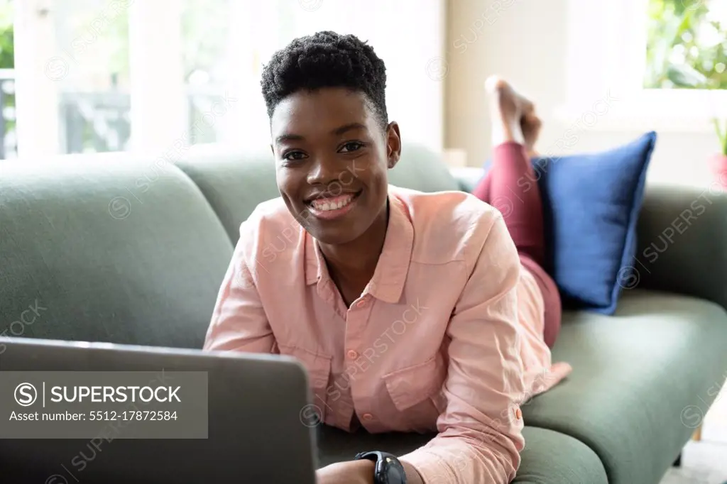 Portrait of african american woman lying on couch using laptop working from home. looking at camera and smiling. staying at home in self isolation during quarantine lockdown.