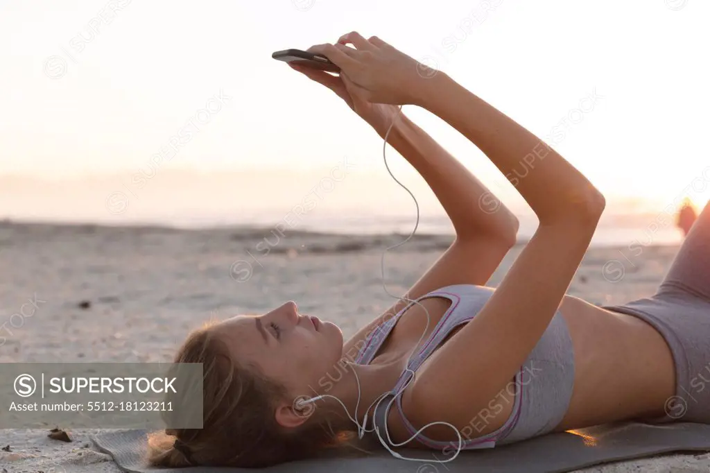 Caucasian woman wearing earphones using smartphone while lying on yoga mat at the beach. fitness yoga and healthy lifestyle concept