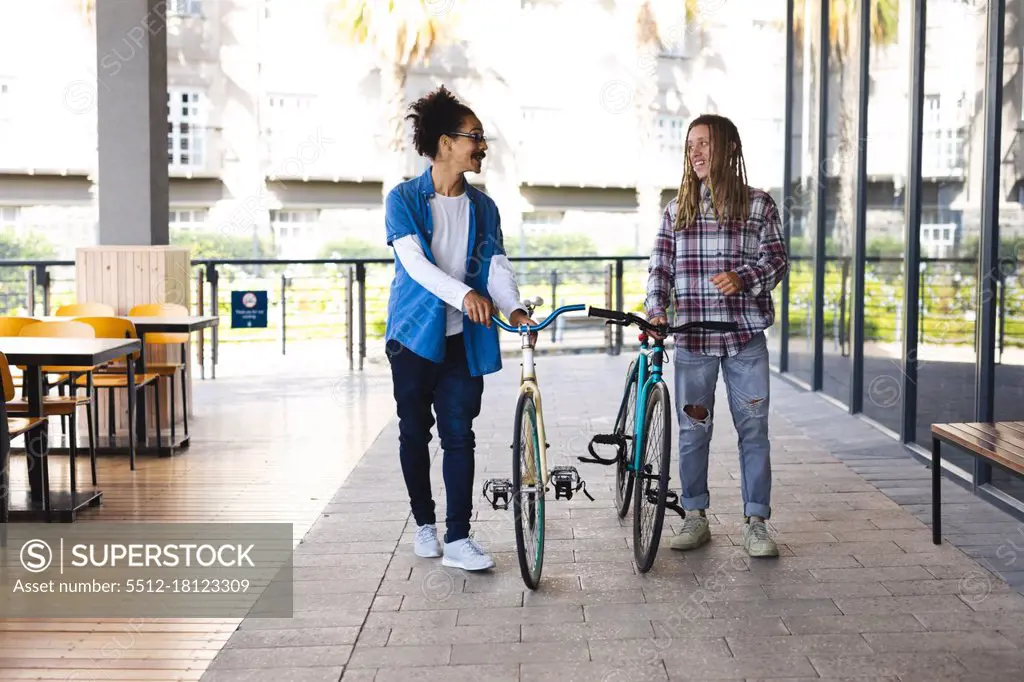 Two happy mixed race male friends wheeling bicycles in the street and talking. green urban lifestyle, out and about in the city.