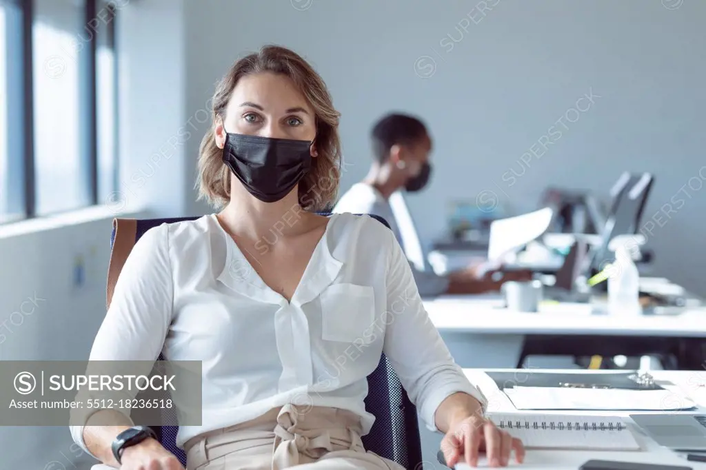 Caucasian businesswoman wearing face mask, sitting at desk, looking at camera. independent creative business at a modern office during coronavirus covid 19 pandemic.