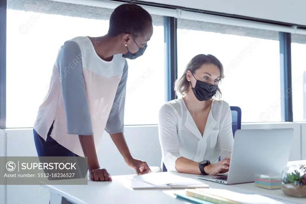 Two diverse smiling businesswomen wearing face masks, working together, using laptop, talking. independent creative business at a modern office during coronavirus covid 19 pandemic.