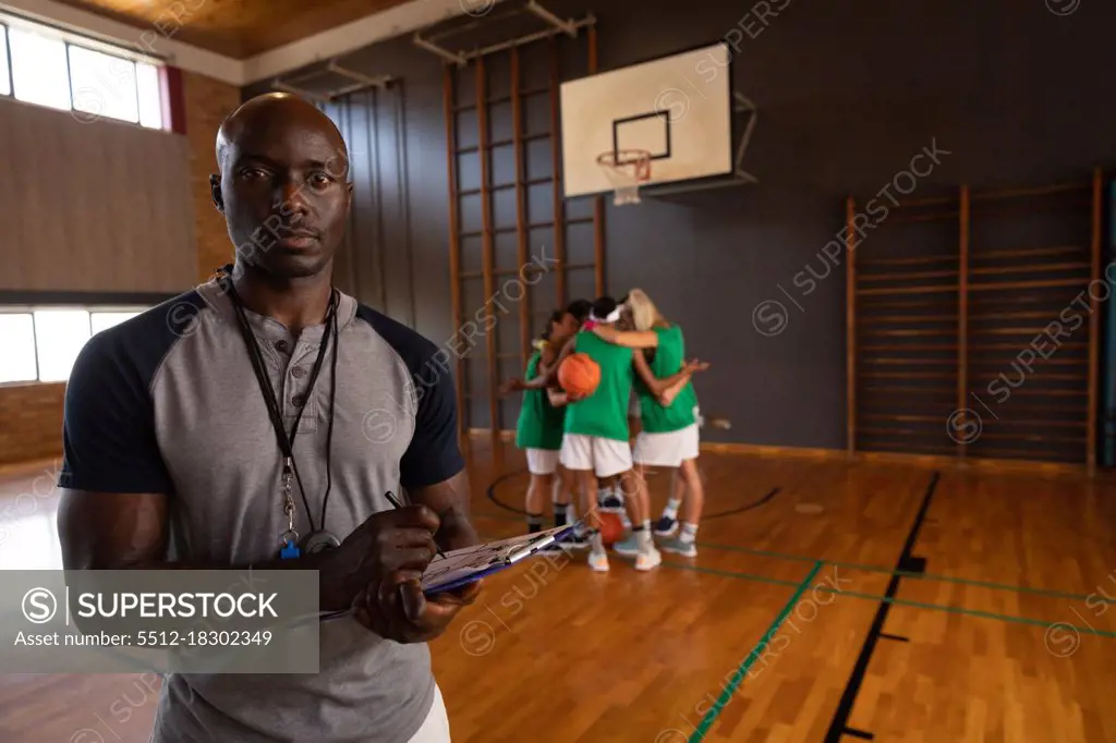 Portrait of african american male basketball coach holding clipboard with team in background. basketball, sports training at an indoor court.