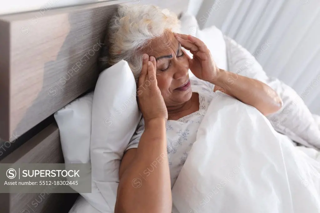 Mixed race senior woman lying in bed holding her head in thought. staying at home in isolation during quarantine lockdown.