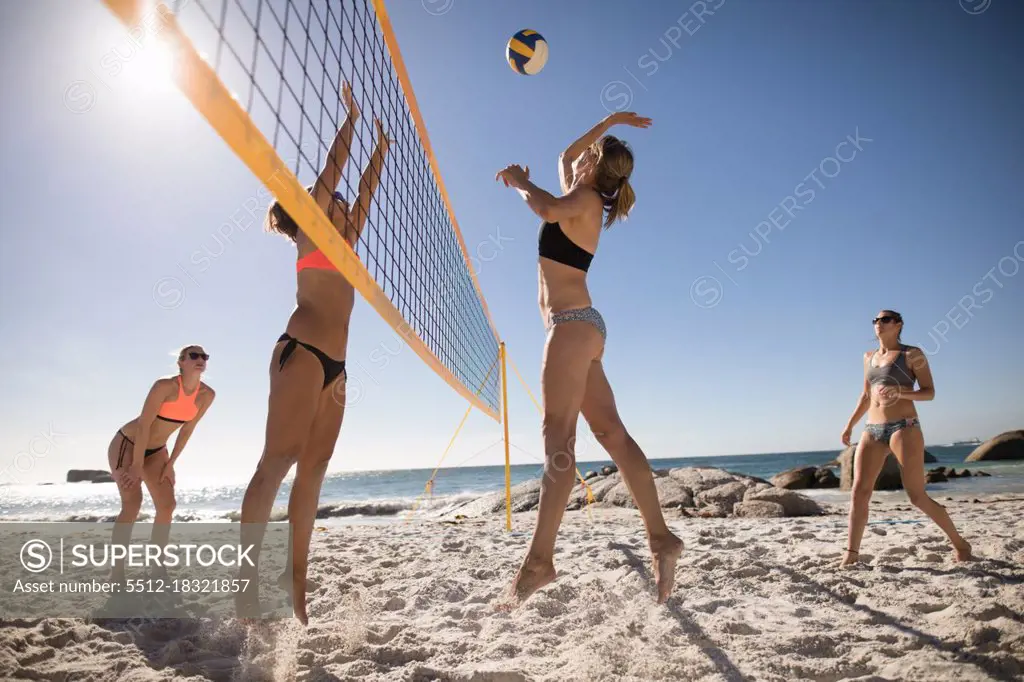 Low angle side view of a group of Caucasian female friends enjoying free time on a beach on a sunny day with blue sky, playing volleyball, hitting a ball