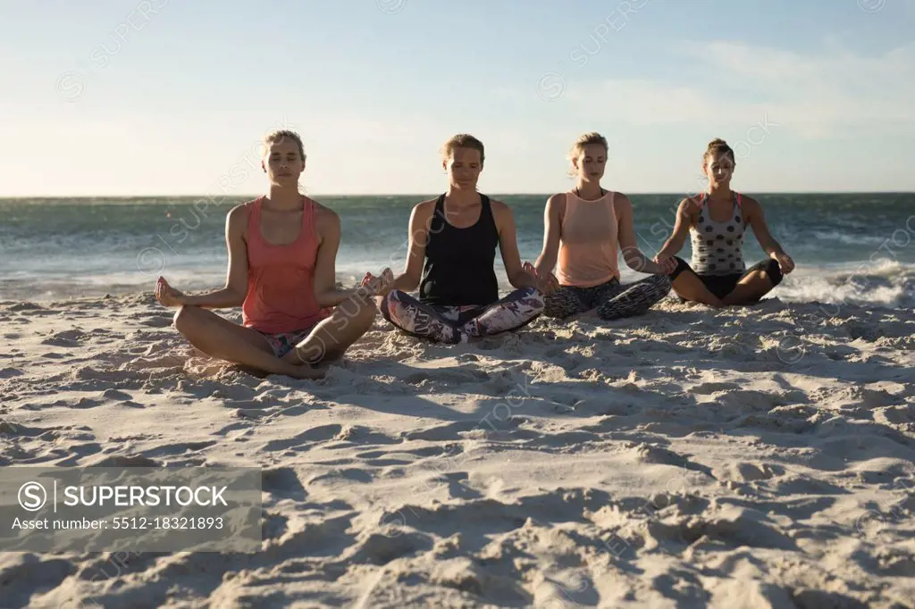 Front view of a group of Caucasian female friends enjoying free time on a beach on a sunny day with blue sky, practicing yoga sitting with legs crossed and eyes closed