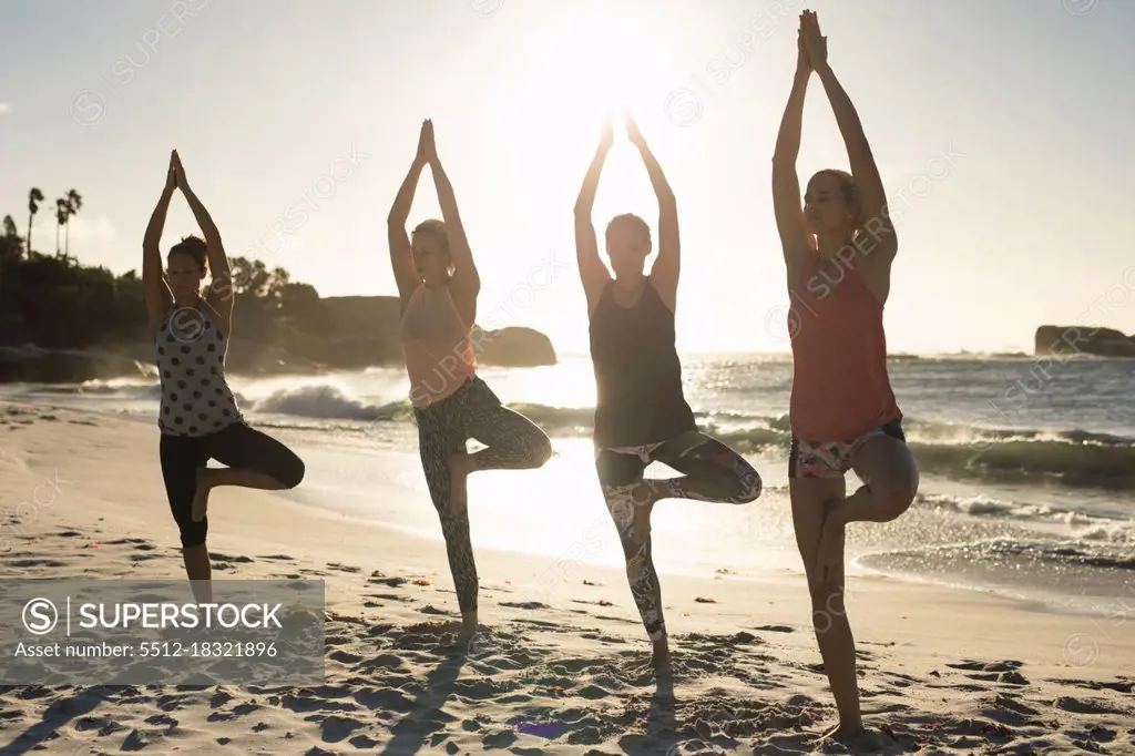 Front view of a group of Caucasian female friends enjoying free time on a beach on a sunny day, practicing yoga 