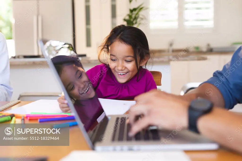 Smiling hispanic daughter sitting in kitchen doing schoolwork with father using laptop in foreground. family spending time together at home.