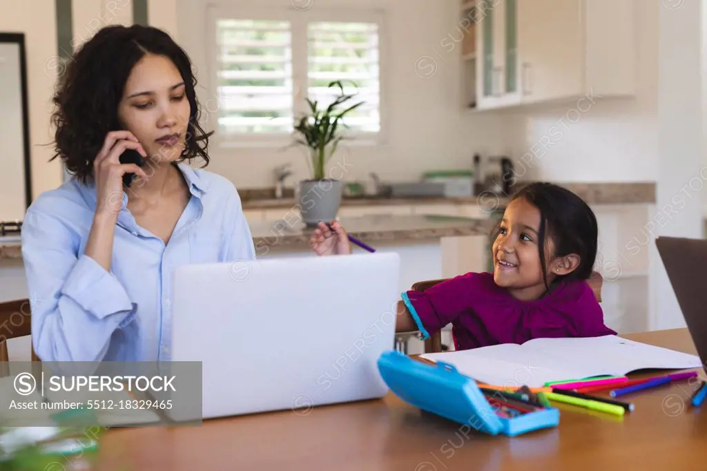 Smiling hispanic daughter in kitchen doing schoolwork with mother using smartphone and laptop. family spending time together at home.