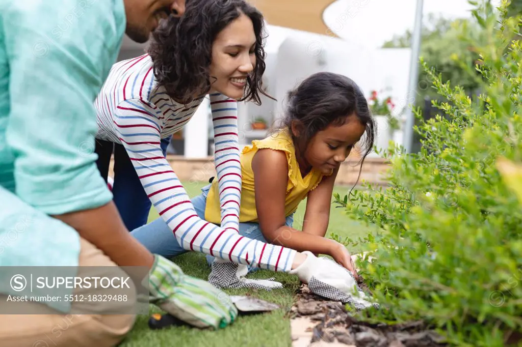 Smiling hispanic mother, father and daughter gardening, kneeling and planting in flower bed. family spending time together at home.