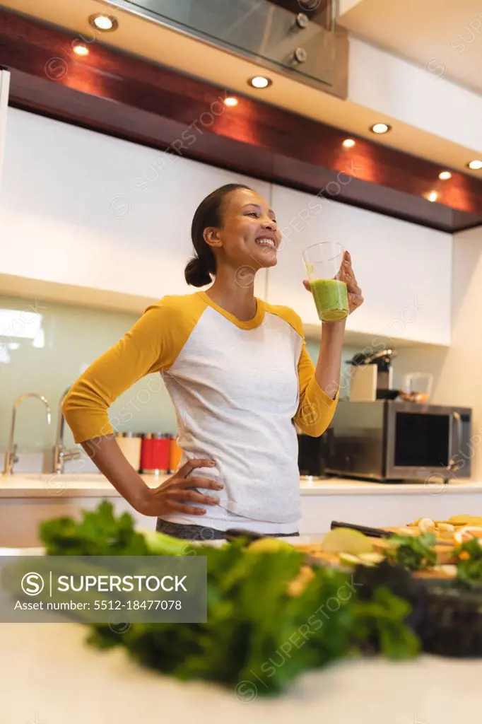 Smiling mixed race woman in kitchen drinking health drink. domestic lifestyle, enjoying leisure time at home.