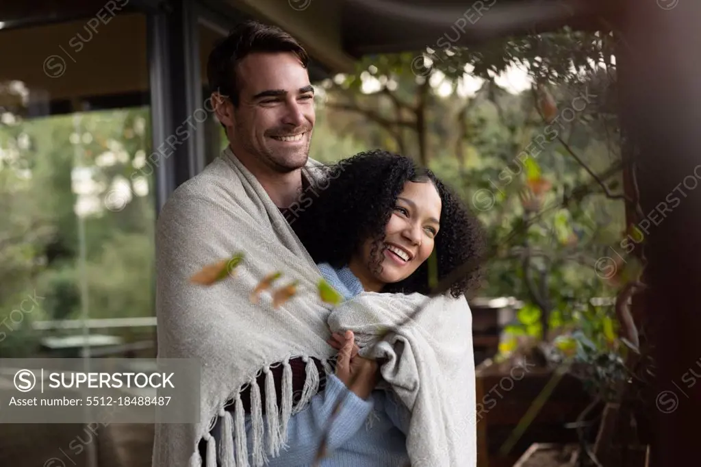 Happy diverse couple covered with blanket on balcony embracing and smiling. spending time off at home.