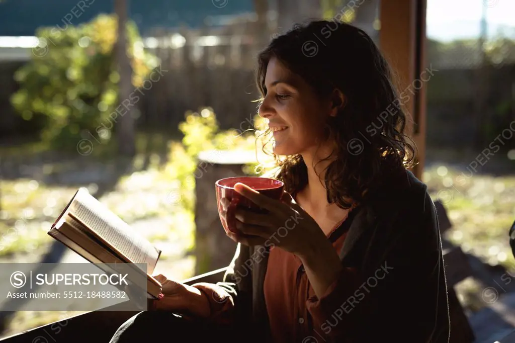 Mixed race woman reading book and drinking coffee in sunny garden. healthy lifestyle, enjoying leisure time at home.