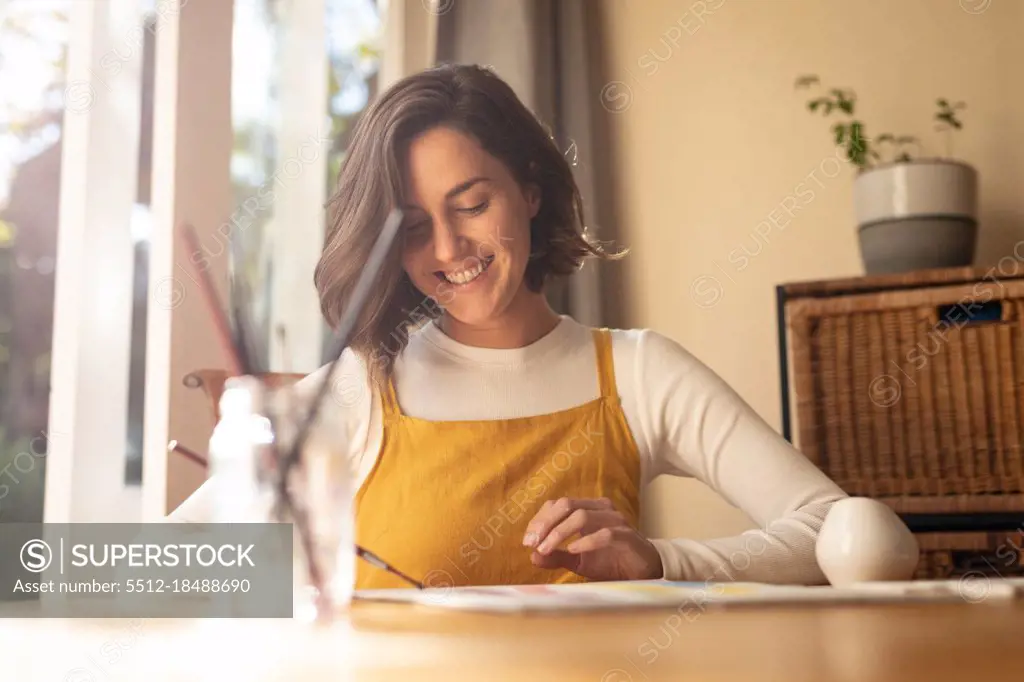 Smiling caucasian woman in living room, sitting at table painting. domestic lifestyle, enjoying leisure time at home.