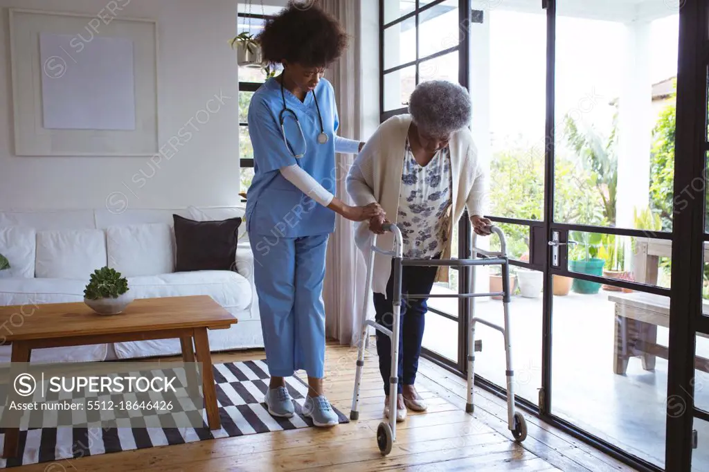 African american female doctor helping senior female patient with walking frame at home. healthcare and lifestyle during covid 19 pandemic.