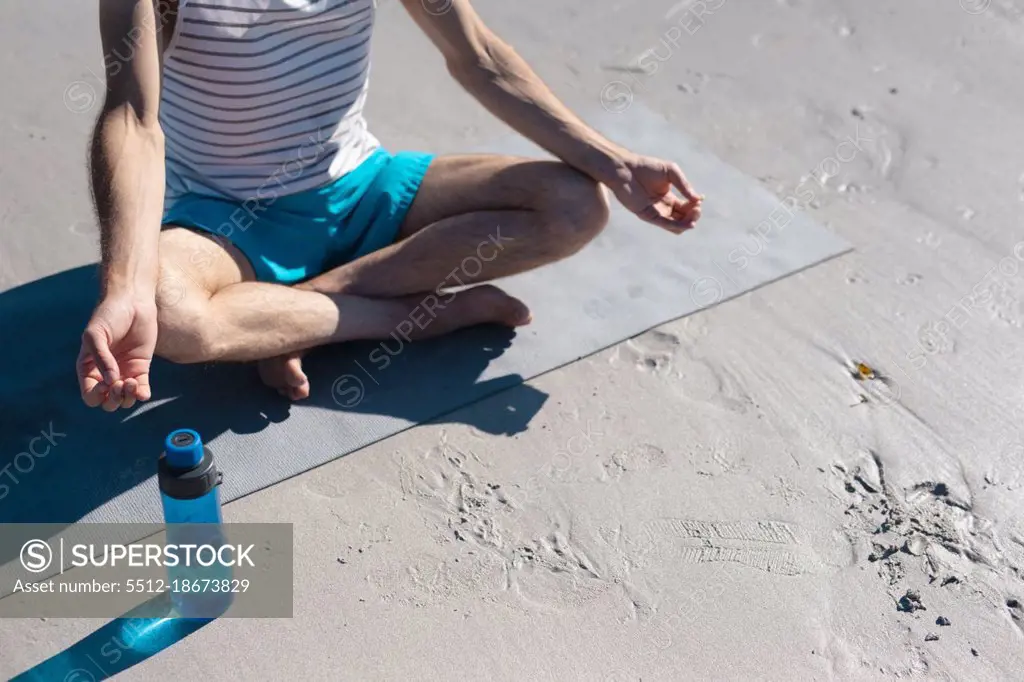 Low section of man meditating while practicing yoga by water bottle on sand at beach. fitness and healthy lifestyle.