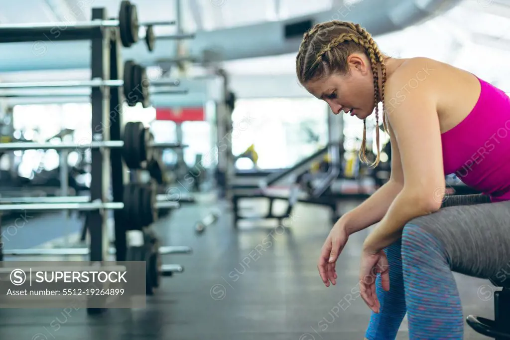 Side view of thoughtful young Caucasian female athletic relaxing on a bench in fitness center. Bright modern gym with fit healthy people working out and training