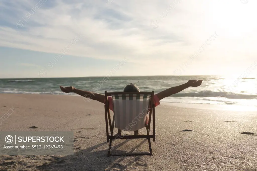 Retired senior biracial man with arms outstretched sitting on folding chair at beach during sunset. lifestyle and weekend.