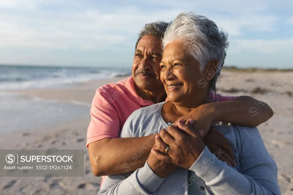 Thoughtful senior multiracial couple embracing while looking away at beach during sunset. lifestyle, love and weekend.