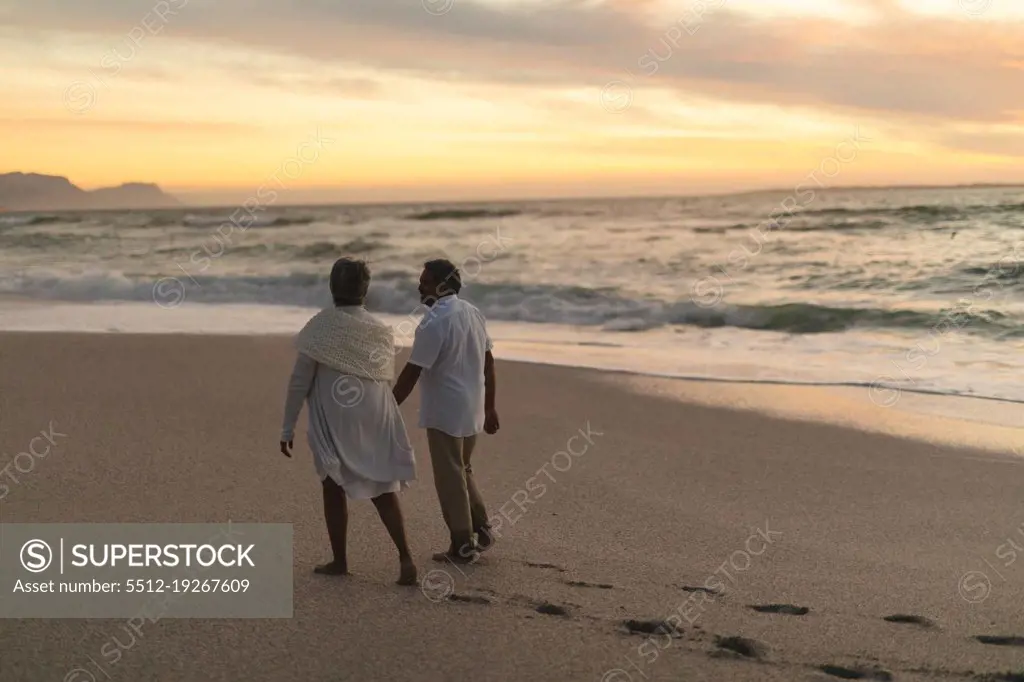 Full length of senior biracial couple walking together on shore at beach during sunset. lifestyle, love and weekend.