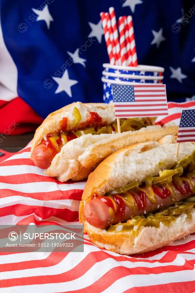 Close-up hot dogs with sauces and jalapenos and american flag, straw and disposable cups on table. unaltered, unhealthy food, meat, bread, sausage and fast food.