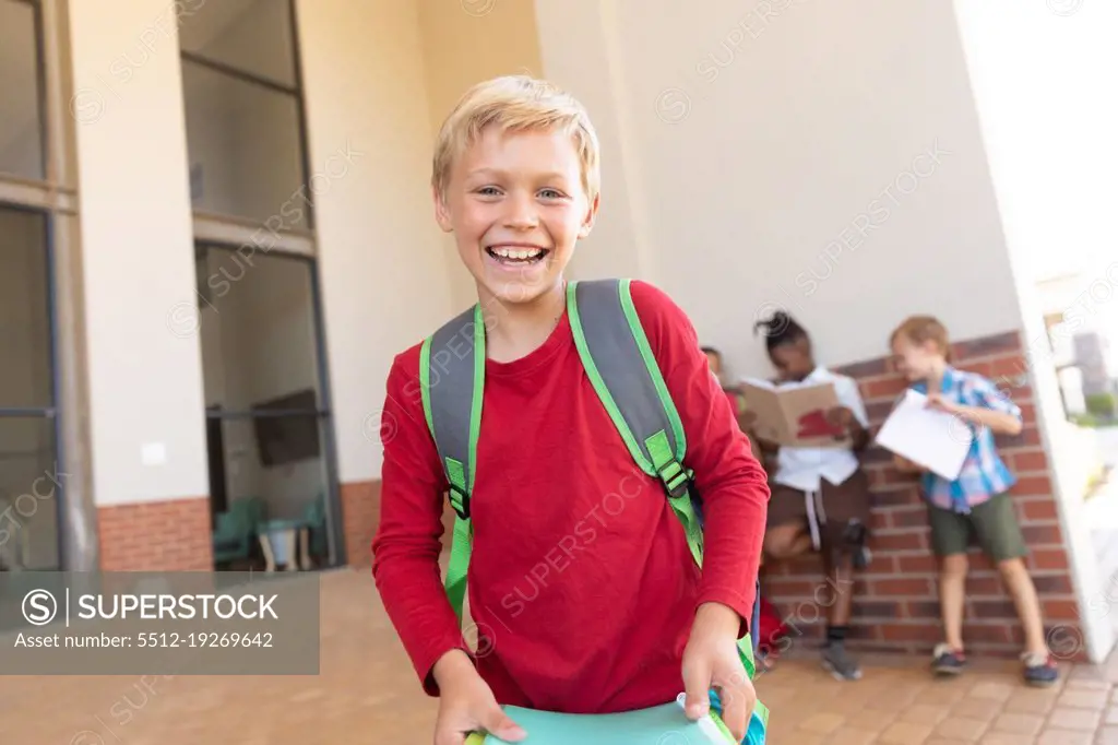 Portrait of cheerful caucasian elementary schoolboy with books standing in school campus. unaltered, childhood, education and back to school concept.