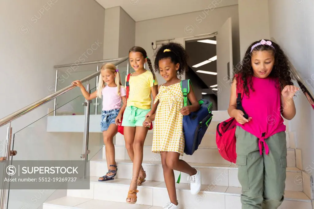 Smiling multiracial elementary schoolgirls with backpacks moving down steps in school building. unaltered, childhood, together, education, enjoyment and back to school concept.