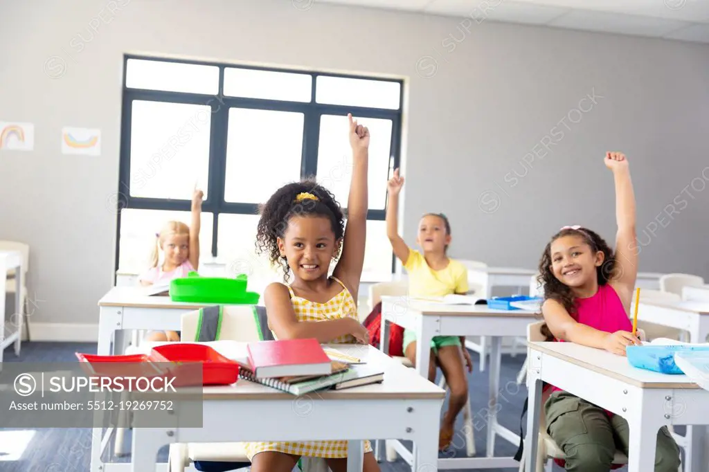 Smiling elementary schoolgirls raising hands while sitting at desk during class in school. unaltered, childhood, education, intelligence and back to school concept.